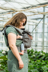 Mom using the Neko Switch Baby Carrier - Bold, 2 extra hoods in a lush greenhouse, supporting her newborn | Babymaxi