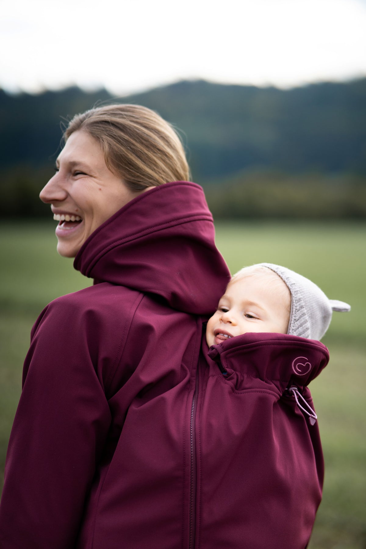 Mamalila Softshell Babywearing Jacket Allrounder Fit Burgundy, laughing mom carrying baby on her back outdoors